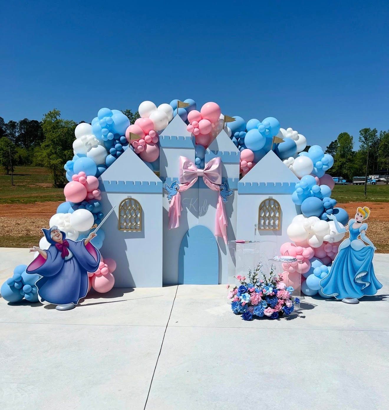 Cinderella-themed party backdrop with castle, balloons, and character cutouts in blue, pink, and white.