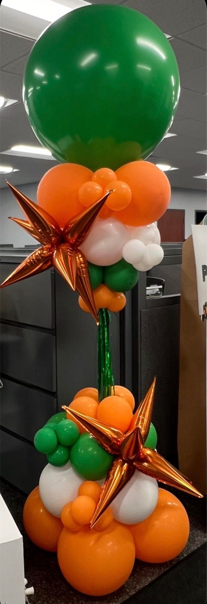Balloon arrangement: green, orange, and white balloons, star accents, standing on a dark surface.