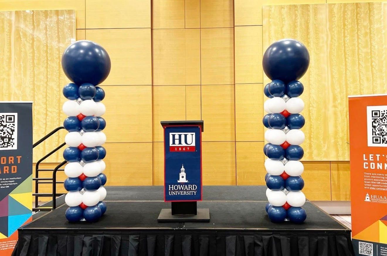 Two balloon columns flanking a podium on a stage. Columns are blue, white, and red. The podium has a logo.
