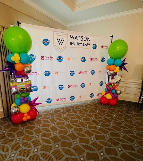 Two balloon columns flanking a backdrop with the Watson Injury Law logo. The columns have a colorful 80s theme.
