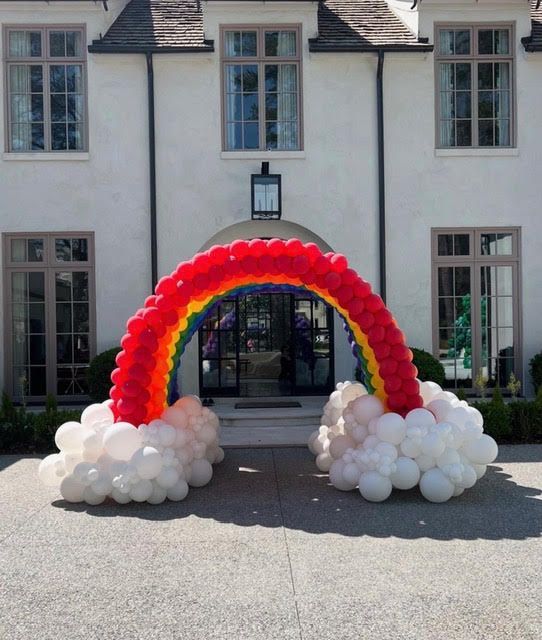 Rainbow balloon archway with white cloud bases in front of a white house with brown trim.
