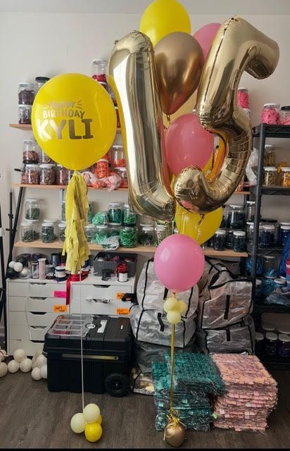 Balloons for a 16th birthday celebration: gold foil numbers, pink and yellow balloons, in a shop setting.