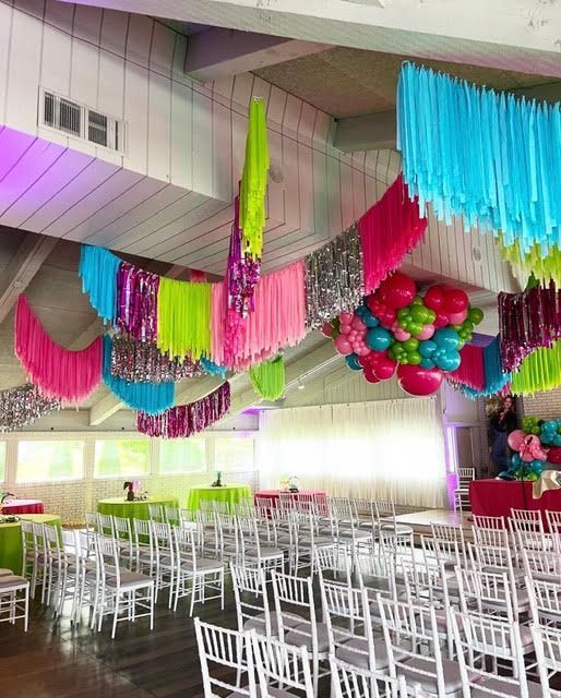 Festive party decor with colorful streamers, balloons, and white chairs set up in an event space.
