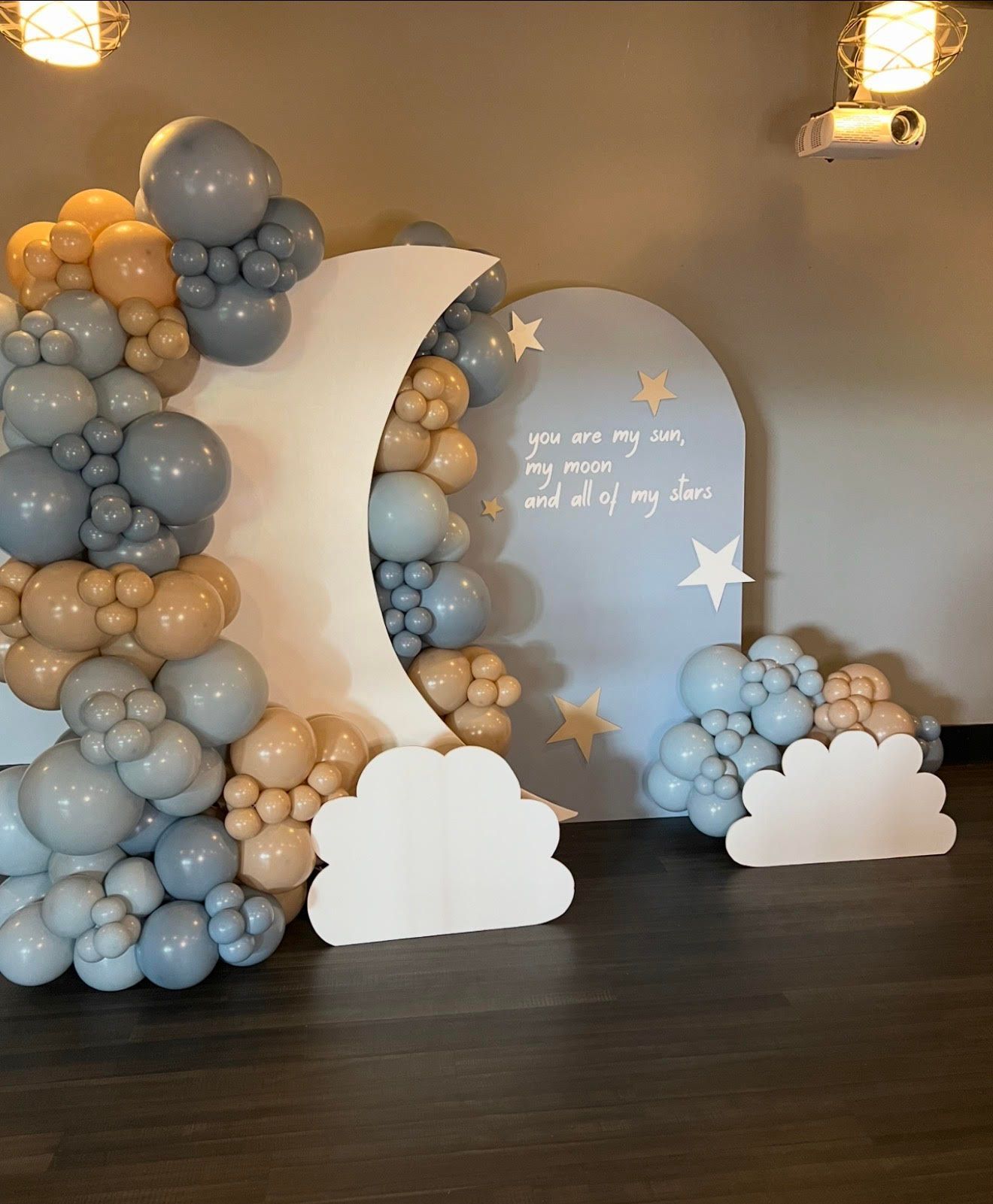 Baby shower backdrop with moon, clouds, stars, and balloon garland in blue, tan, and white.