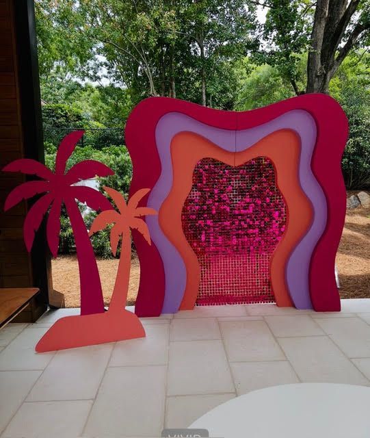 Colorful layered backdrop with pink sequin center, two palm tree cutouts, outdoors.