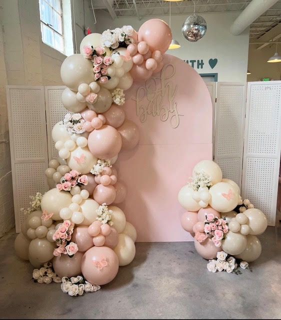 Pink balloon arch with flowers against a light pink backdrop, celebrating a party.