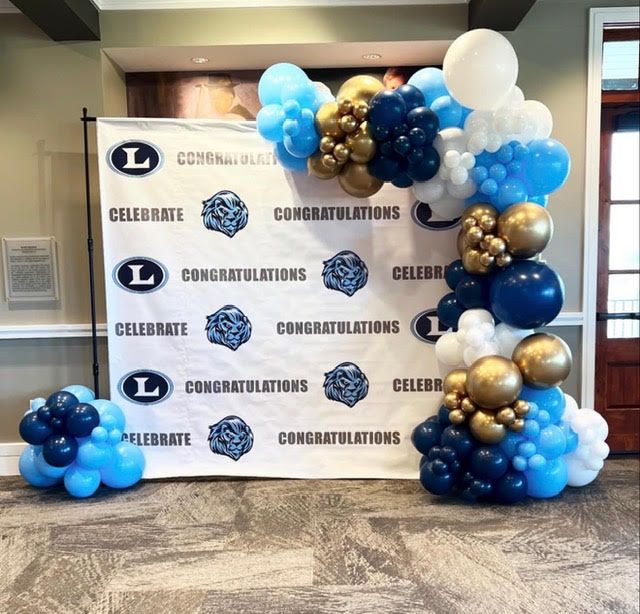 Balloon arch in blue, gold, and white, with matching clusters, by a banner that says 