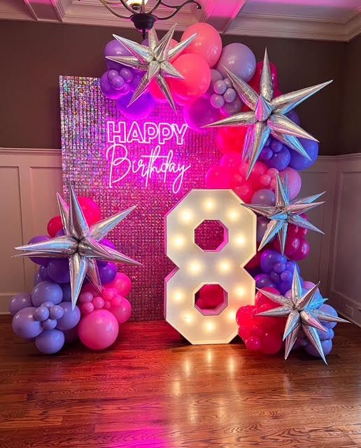Birthday party backdrop with balloons, starburst decorations, neon sign, and a light-up 