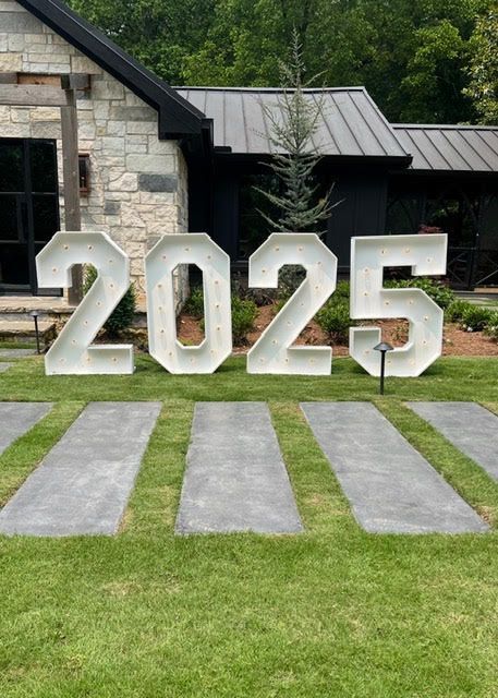 Large white illuminated numbers 2025 on a lawn, next to a modern building with a dark roof.