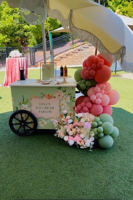 Ice cream cart decorated with floral design and balloons, on a green lawn.
