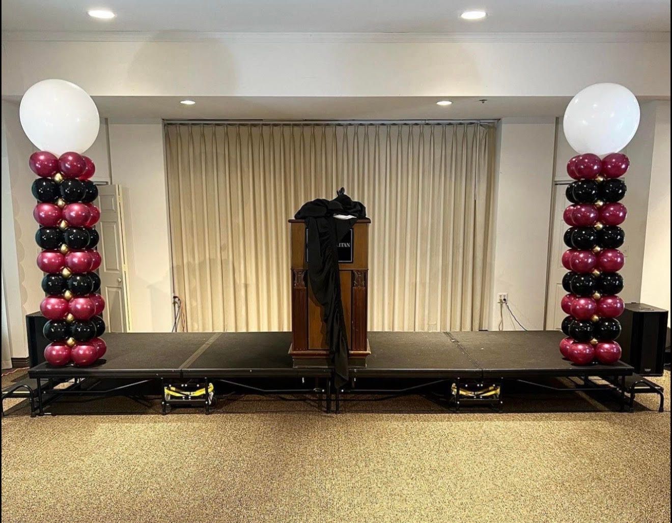 Stage with balloon columns in maroon, black, and white, with a podium draped in black fabric.