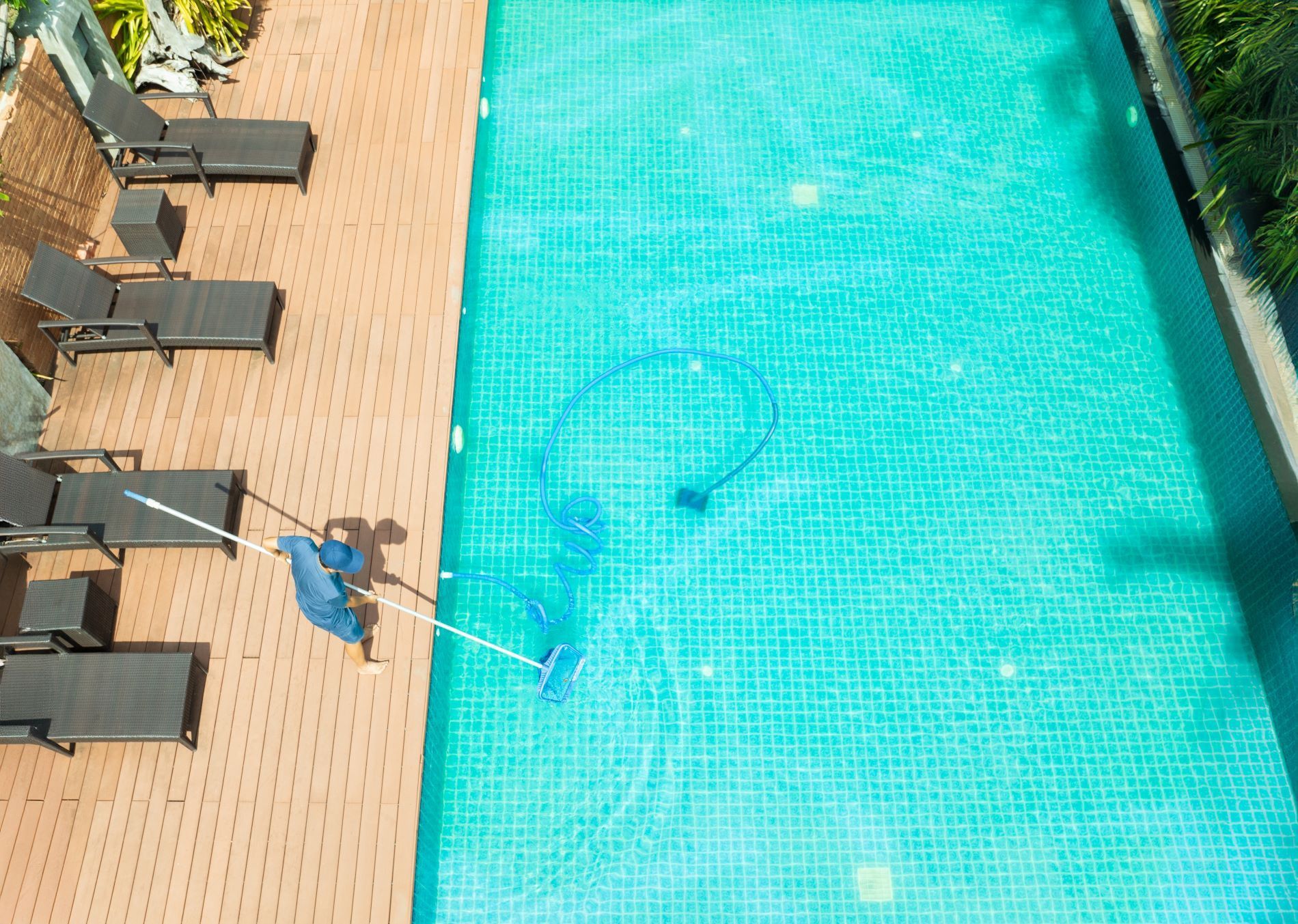 Person cleaning a turquoise tiled swimming pool with a pool vacuum, next to wooden lounge chairs.