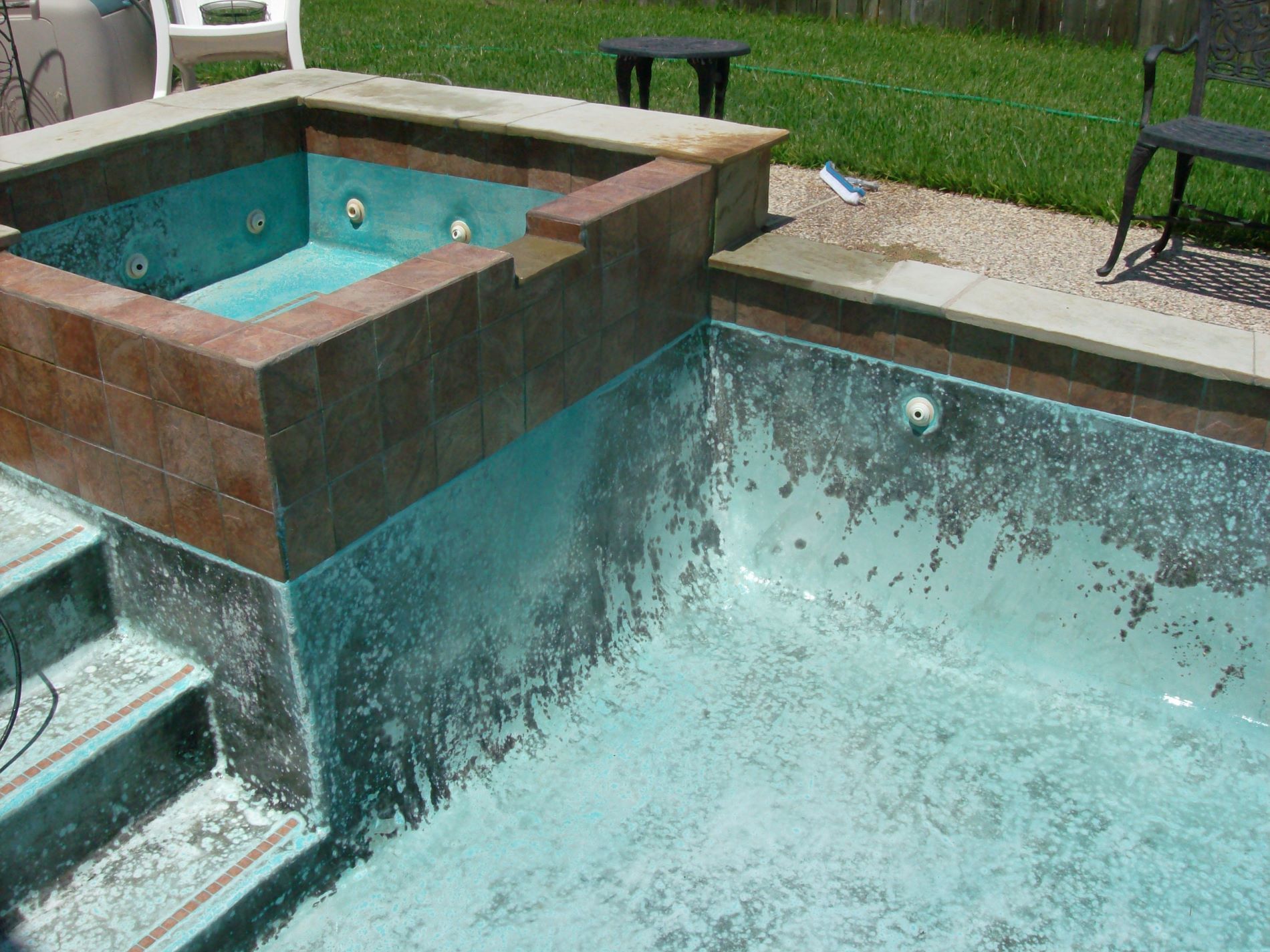 An empty pool and jacuzzi with peeling aqua paint, brown tile and brick detailing, set in a backyard.