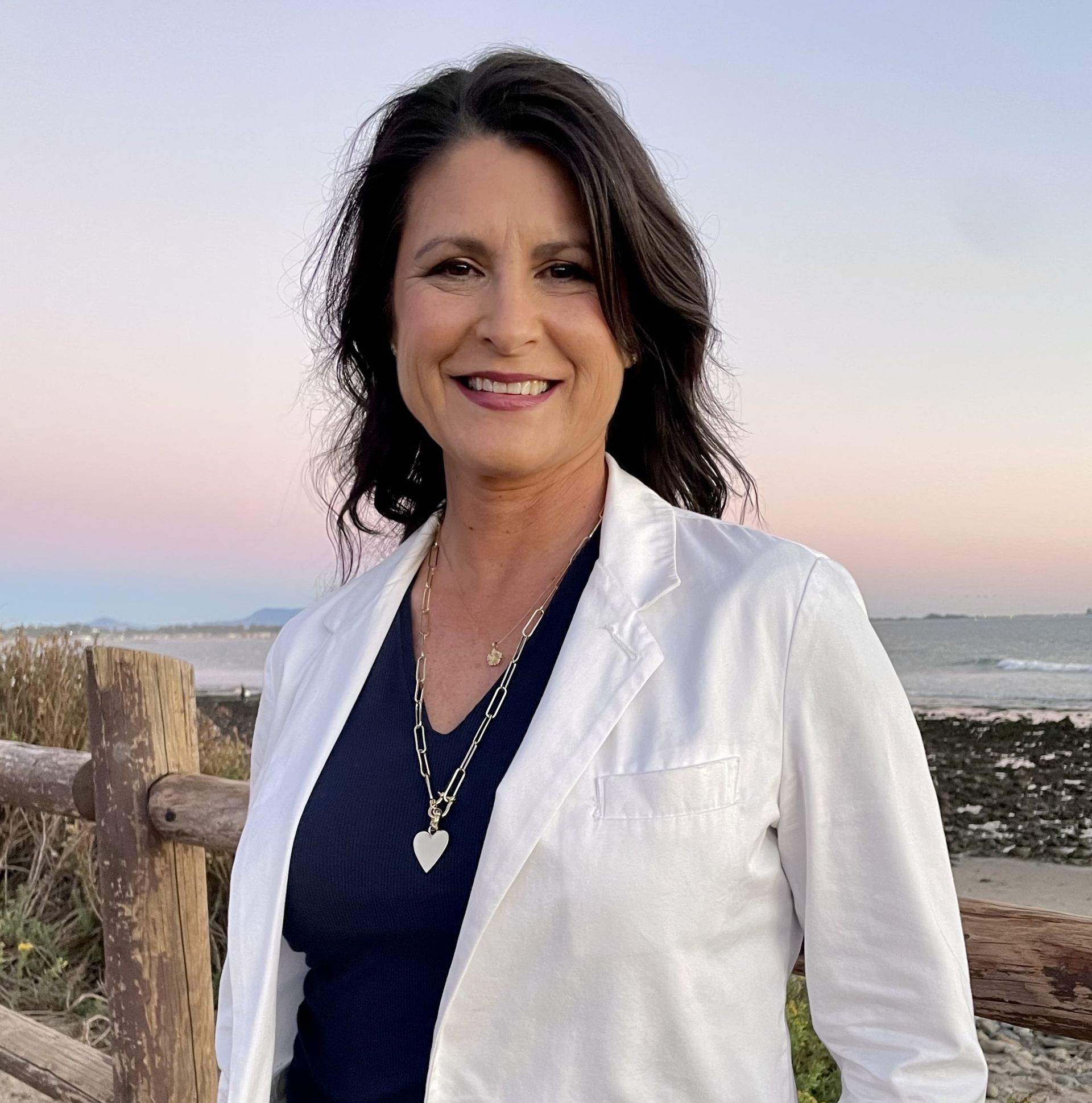 A woman wearing a white jacket and a heart necklace smiles for the camera