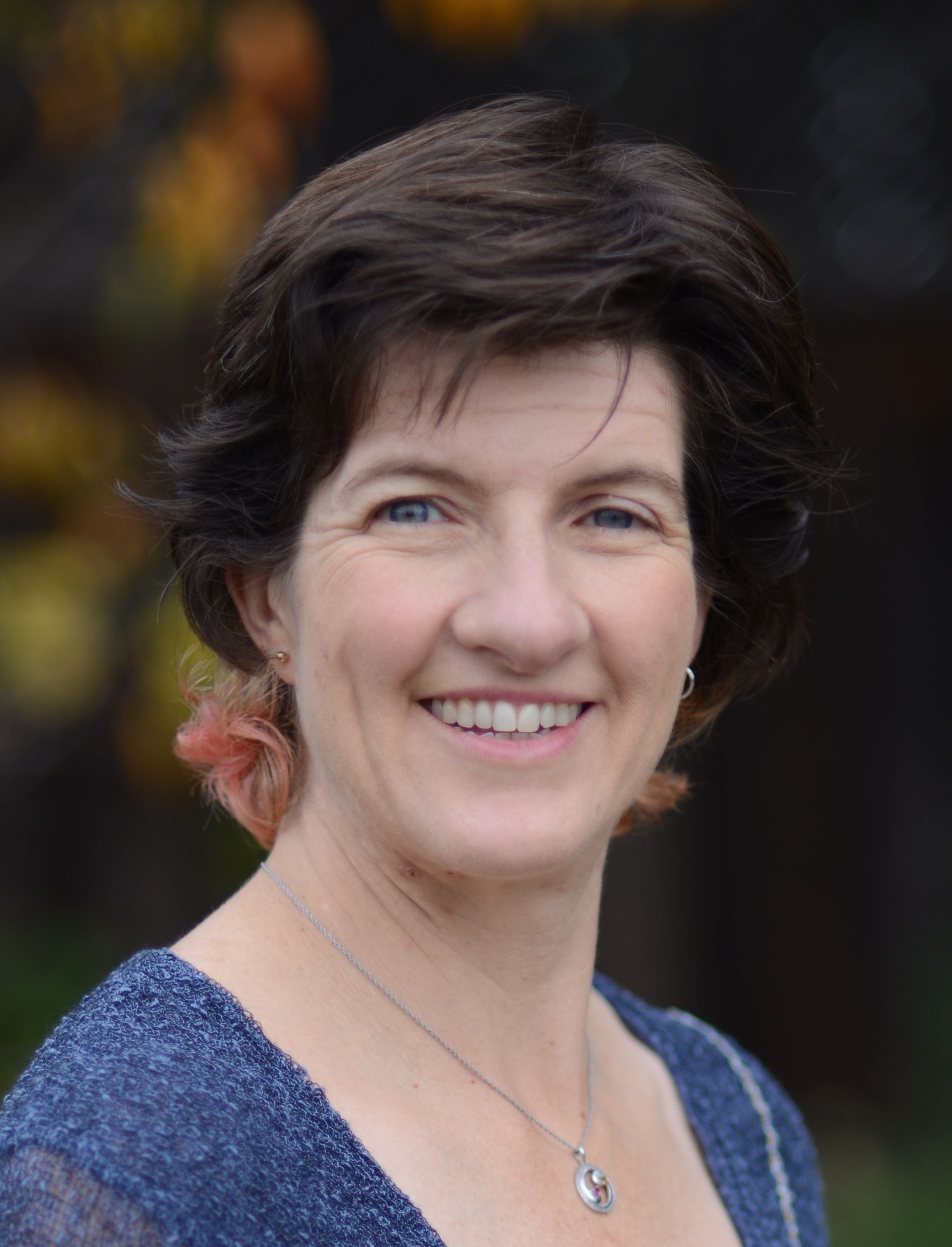 A woman wearing a blue shirt and a necklace is smiling for the camera.