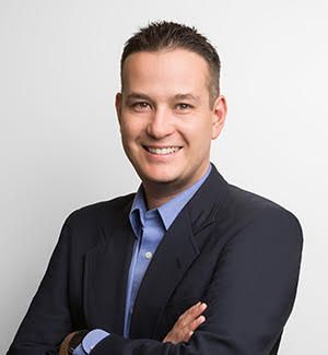 A man in a suit and blue shirt is smiling with his arms crossed.