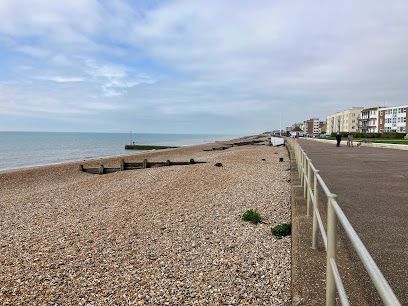 Beach with pebbles, ocean, promenade with railing, buildings along the coast, cloudy sky.