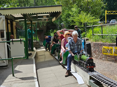 People ride a miniature train at a station. Passengers sit in green cars. Lush greenery surrounds the track.