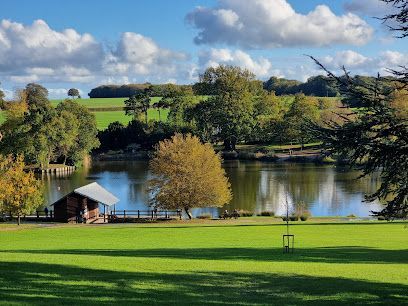 Lush green park with a lake. Small wooden shelter on the lake's edge; trees and rolling hills in the background. Blue sky with clouds.
