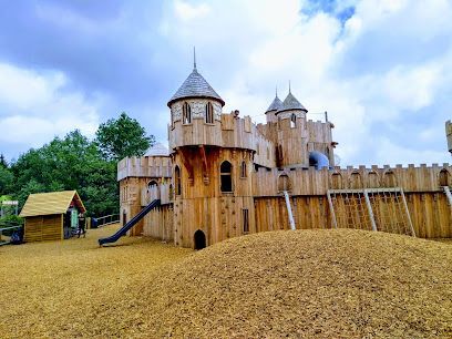 Wooden castle-themed playground with towers, a slide, and a small building under a cloudy sky.