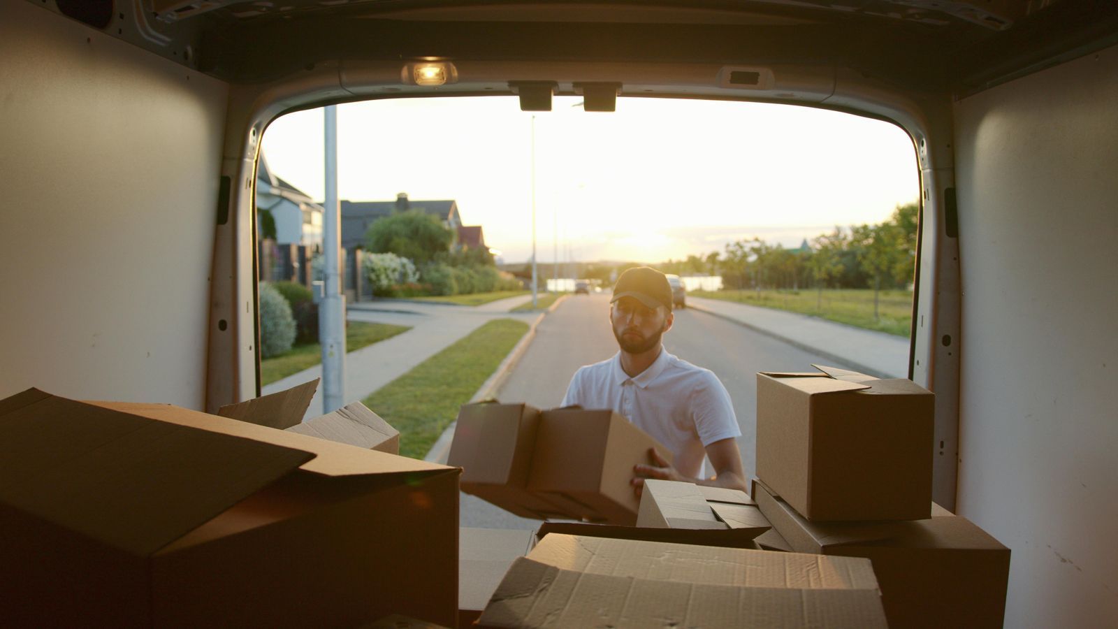 A delivery worker loads cardboard boxes into the back of a van during a sunset in a residential neighborhood.