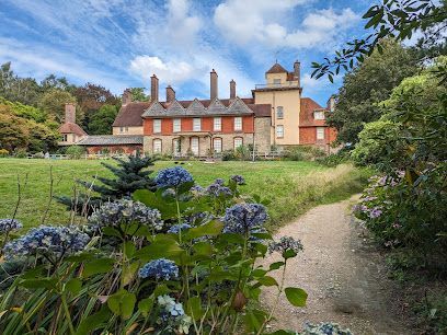 Stone mansion with red brick facade, green lawn, blue hydrangeas, and a gravel path under a cloudy sky.