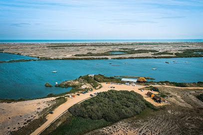 Aerial view of a coastal landscape with a lagoon and ocean. Sandy terrain surrounds a small cluster of buildings and greenery.