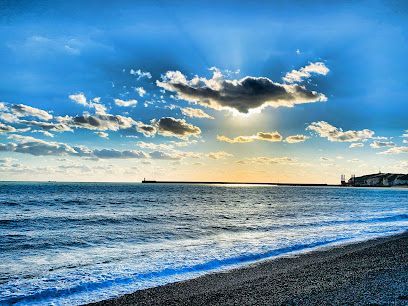 Ocean scene under a bright blue sky with sun breaking through clouds, waves washing on a pebble beach.