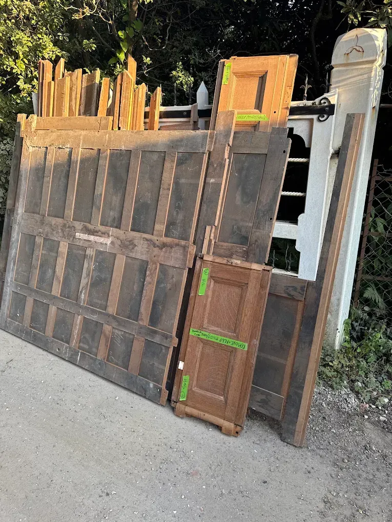 Wooden panels and doors leaning against a white gate on a paved surface, outdoors.