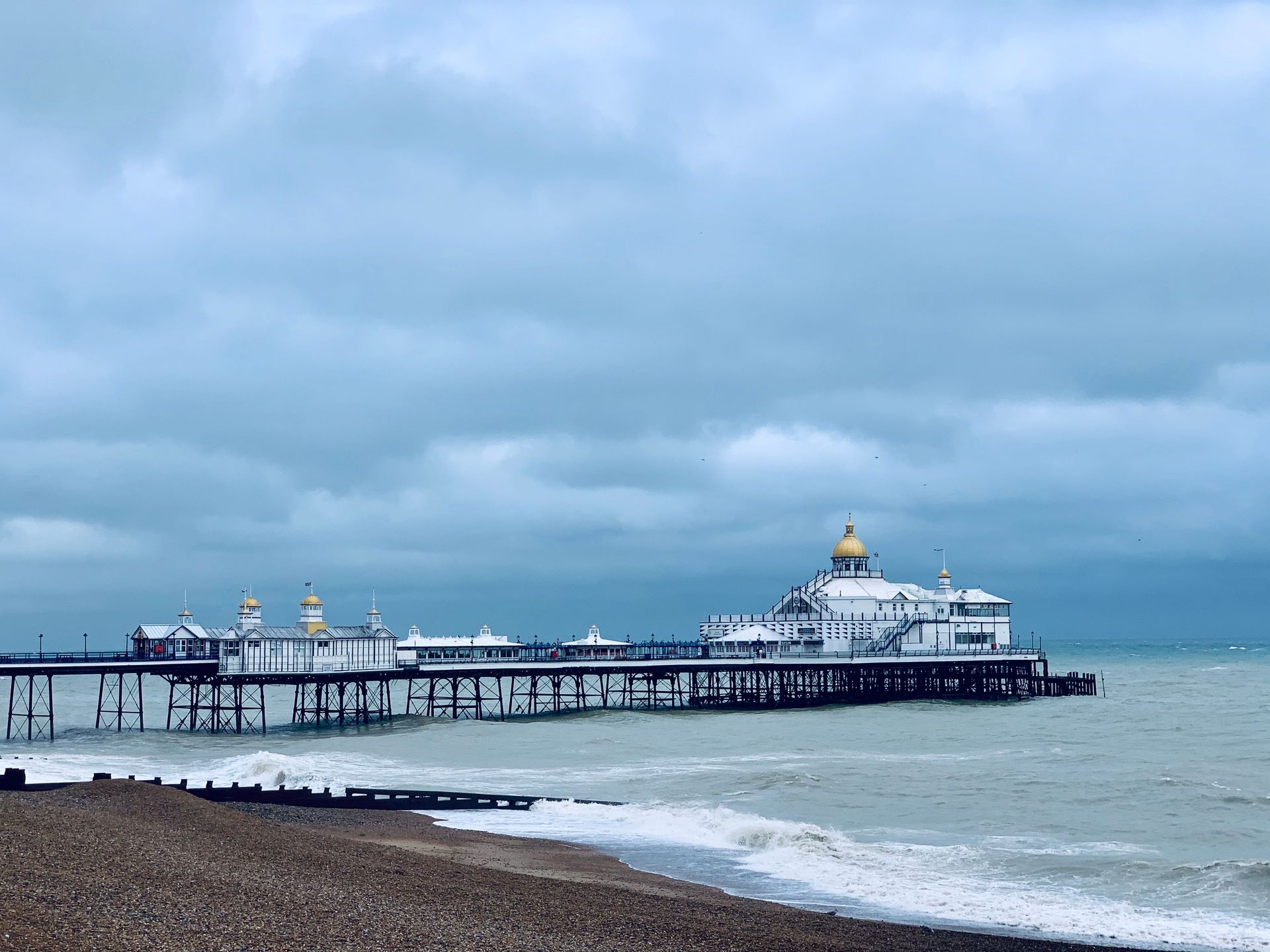 Seaside pier with a white temple-like building under cloudy skies, beside a rocky shoreline and waves