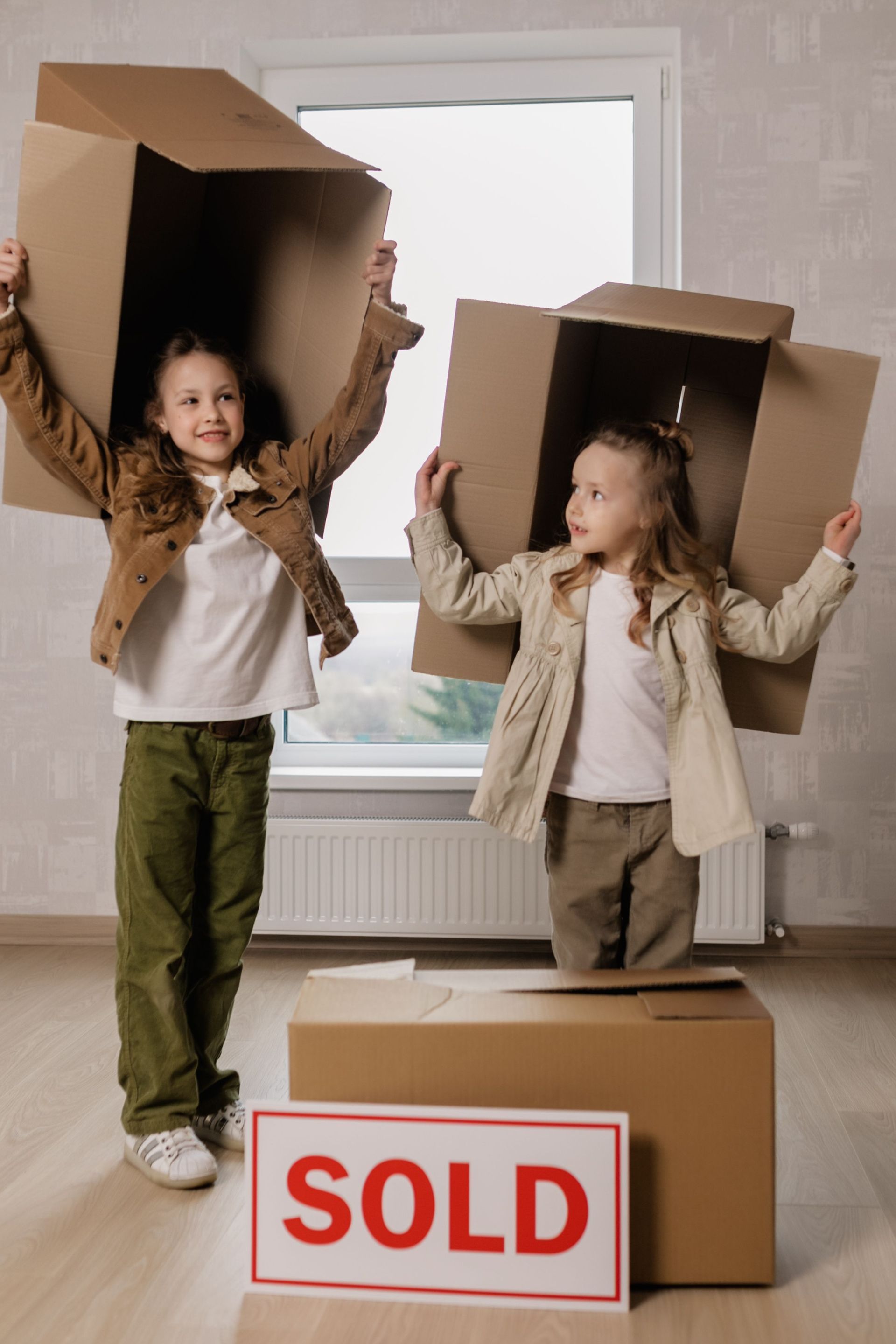 Two children holding cardboard boxes near a box labelled “SOLD” in a bright room