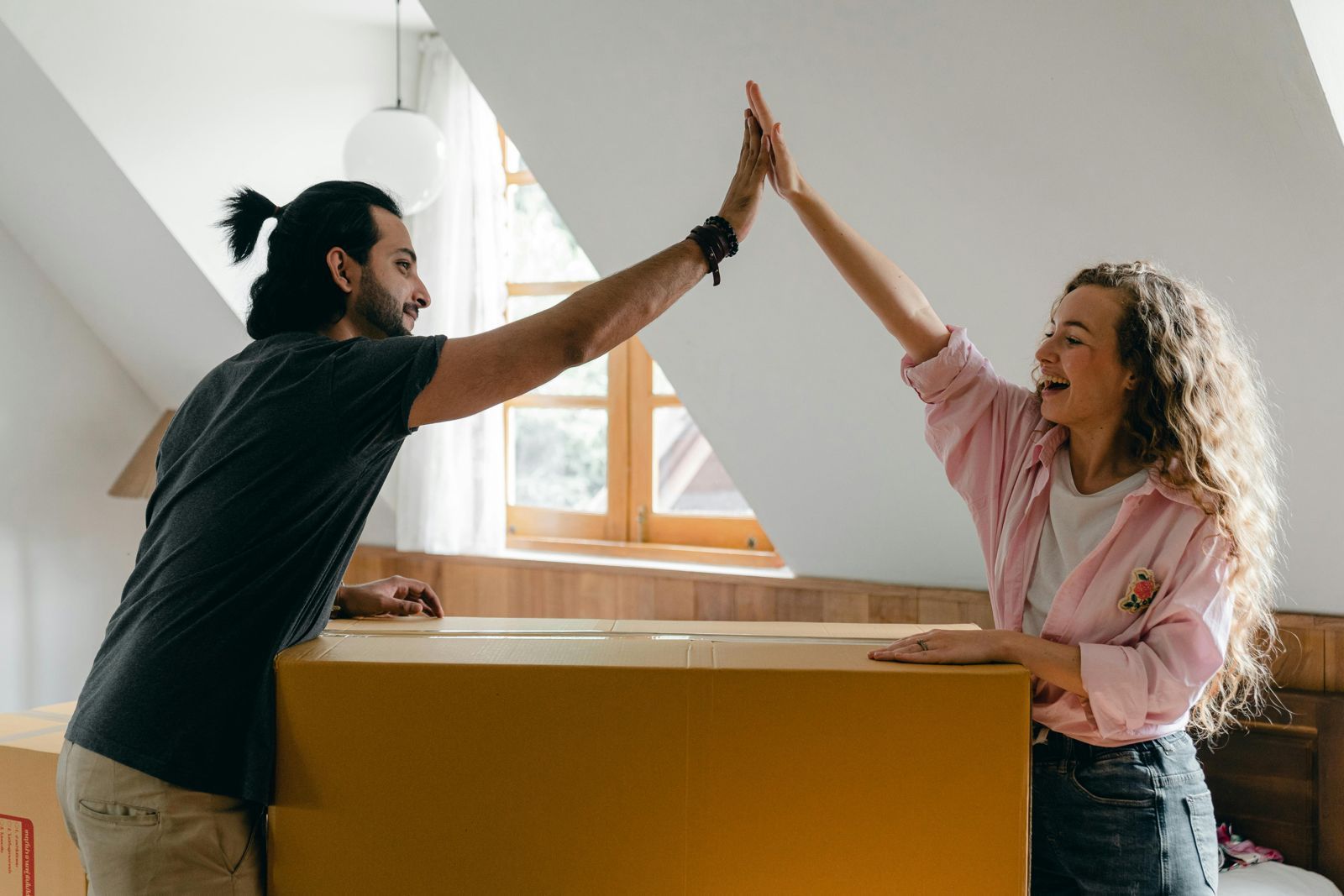 Two people high-five over a large cardboard box in a bright room with a window.