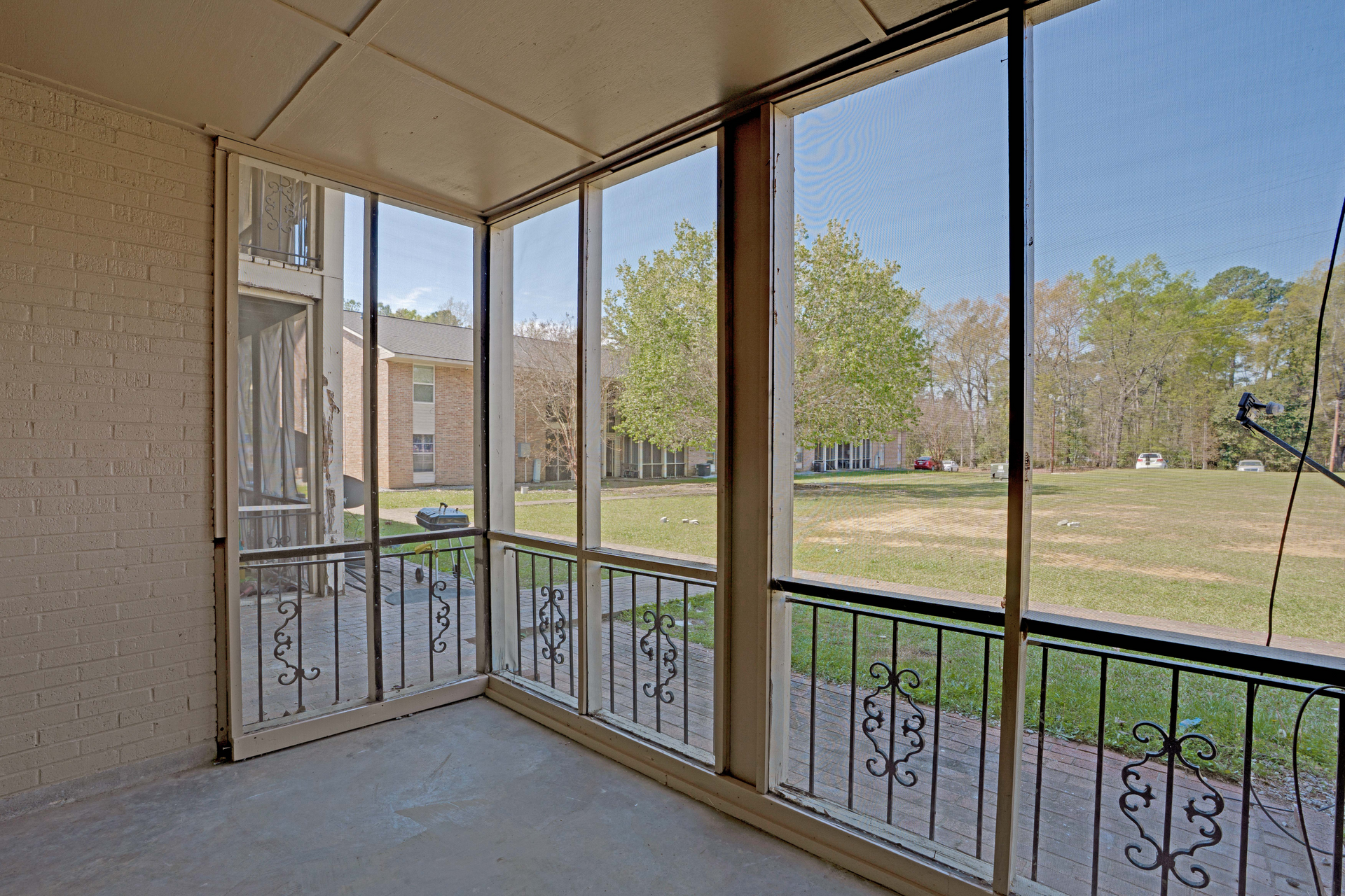 A screened-in porch with light beige walls and decorative metal railings overlooking a grassy lawn and trees.