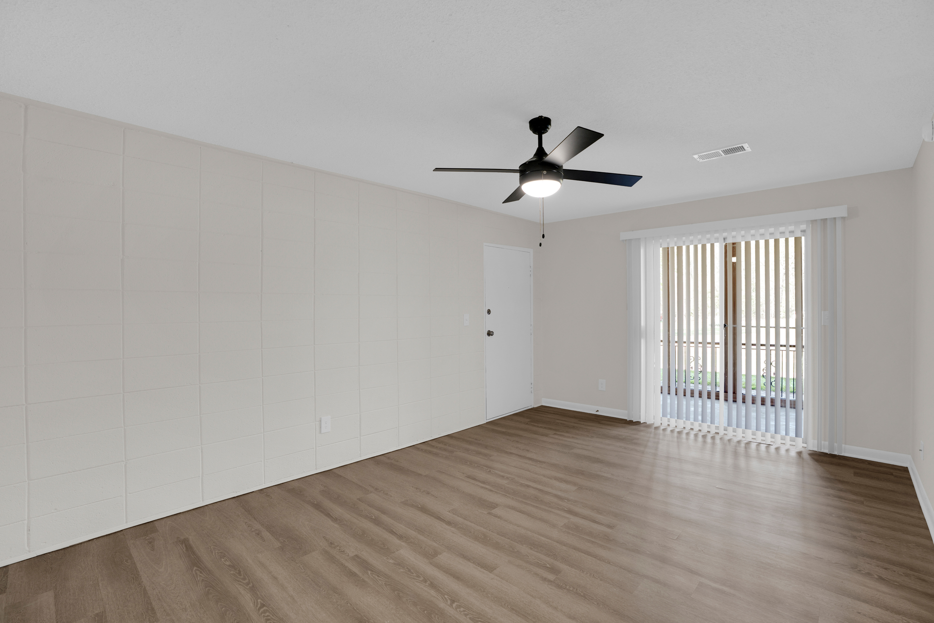 A bright, empty room with light gray walls, wood-look flooring, a ceiling fan, and a large sliding glass door with blinds.