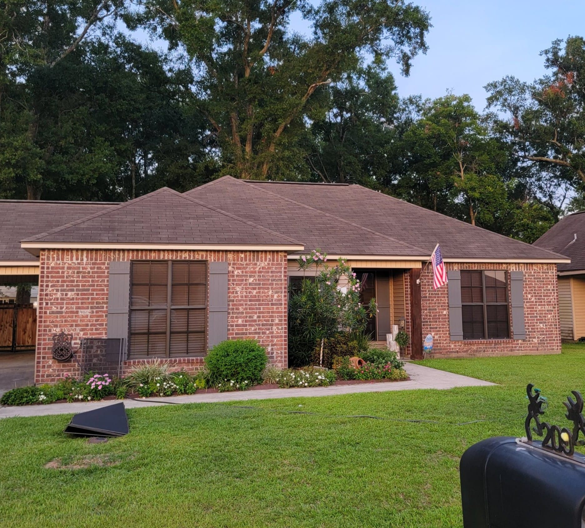 Red brick house with brown roof and green lawn, American flag, trees in background.