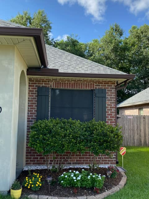 Corner of a brick house with dark green shutters, brown gutters, and a well-manicured flower bed under a blue sky.