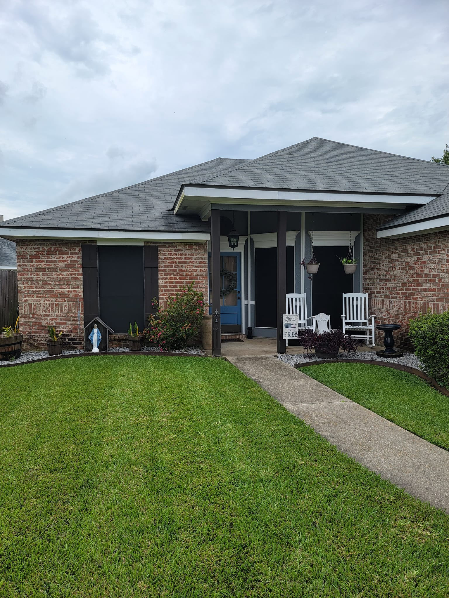 Brick house with dark shutters, front porch with white chairs, green lawn.
