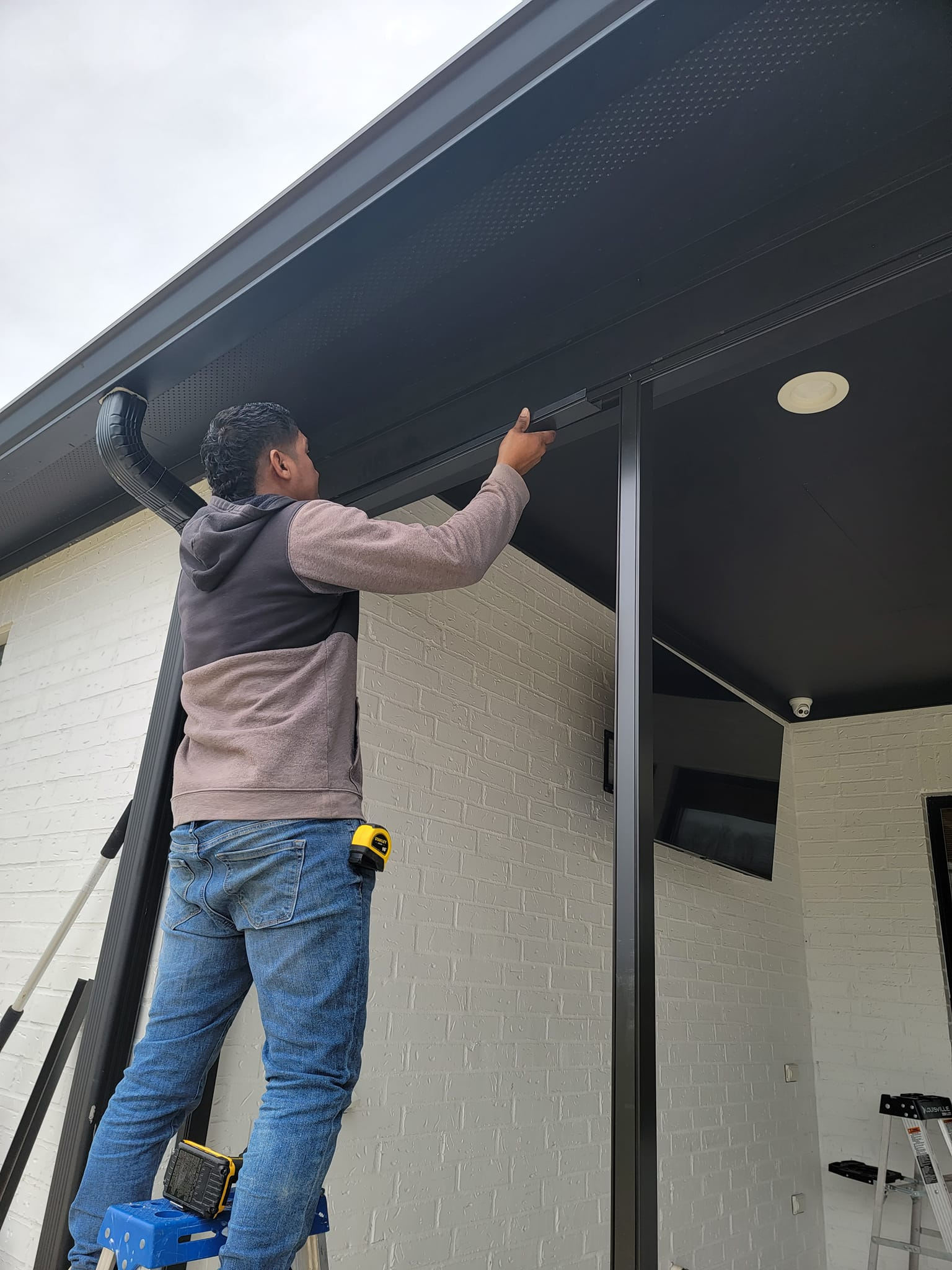 Man on a ladder installing black material on a building's roof underhang; beige wall, gray trim.