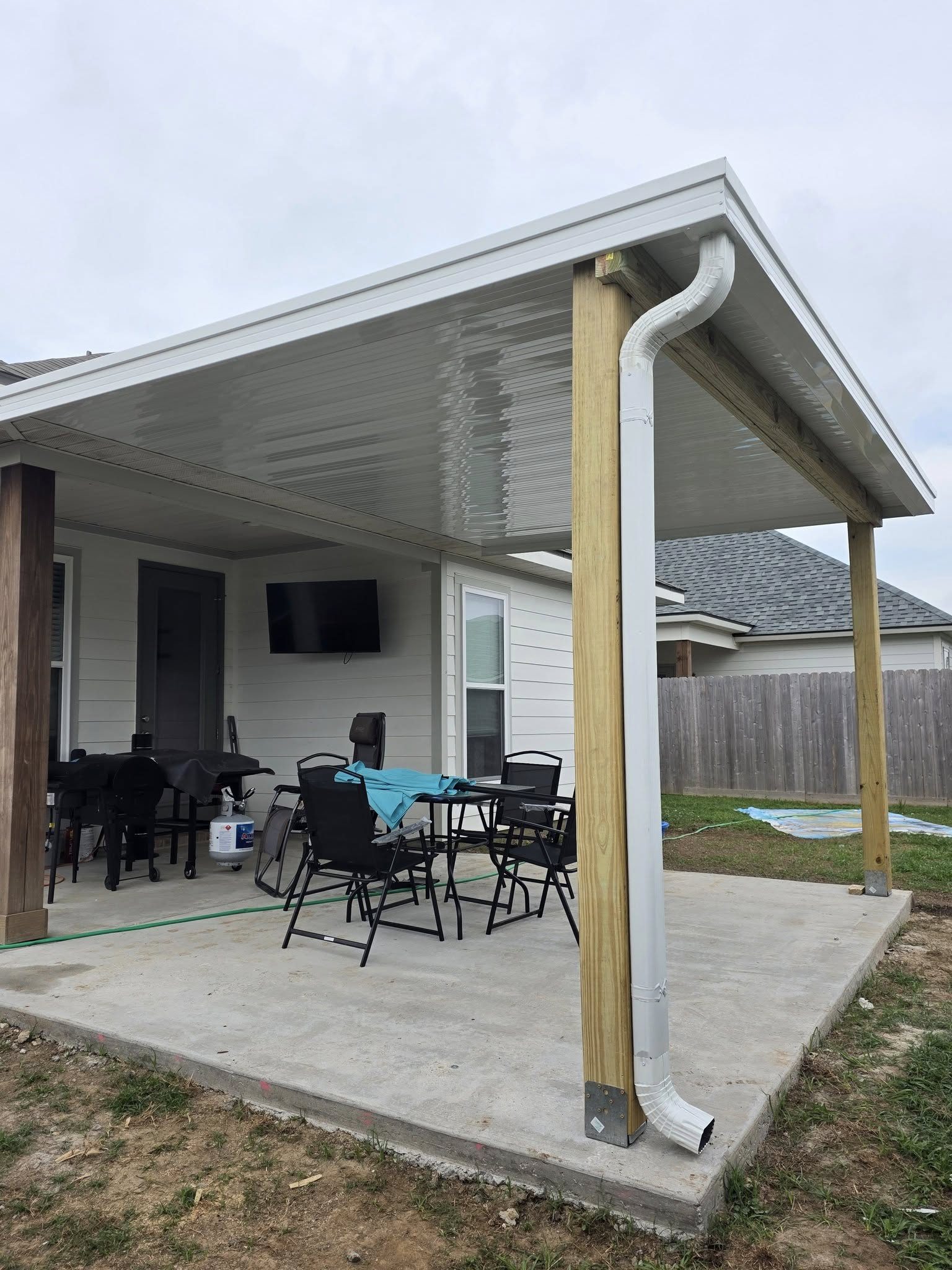 Patio with concrete slab, covered by a white roof supported by wooden posts. Outdoor furniture sits on the patio.