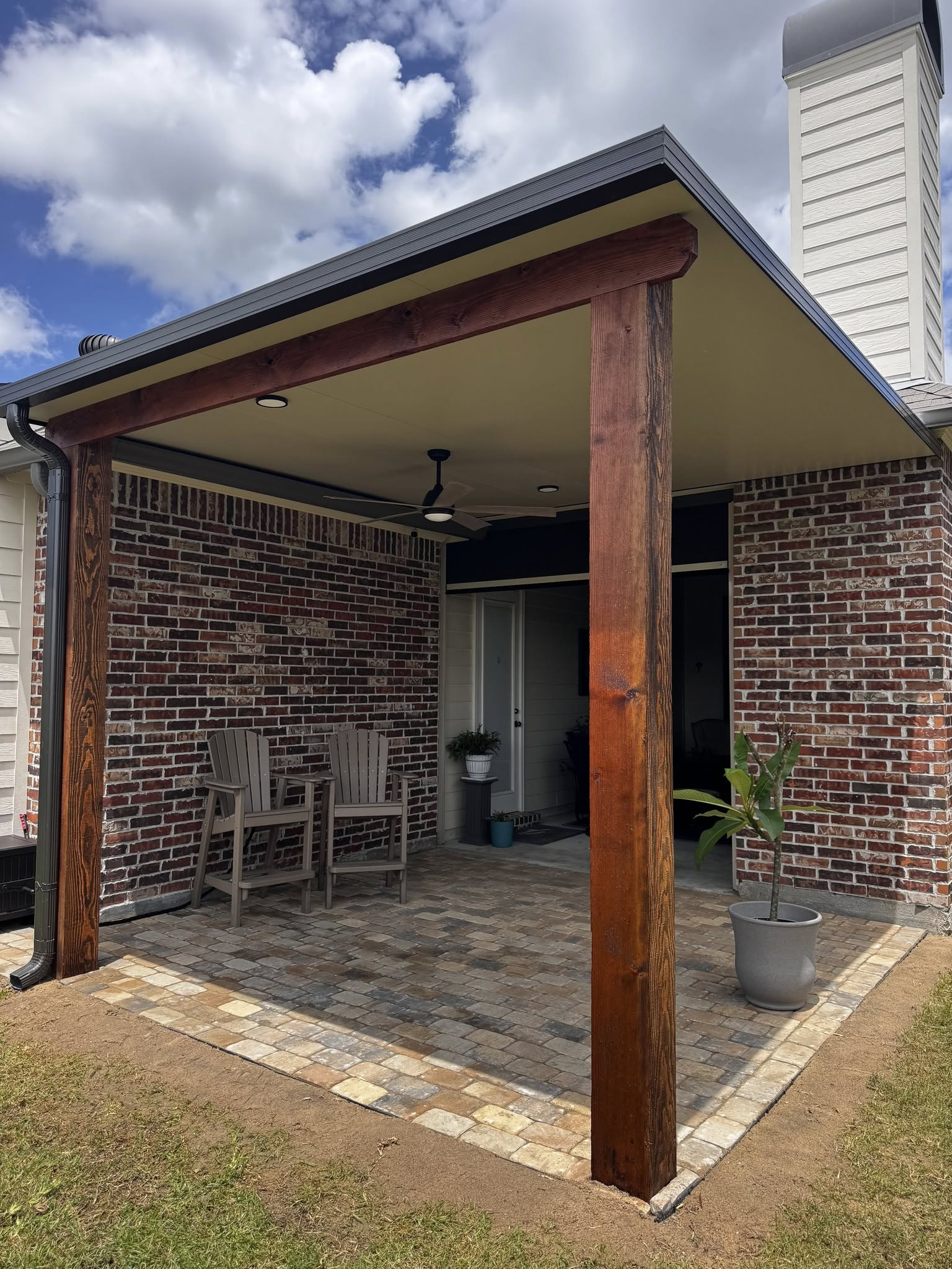 Patio with brick wall, wooden supports, and tiled flooring. A covered seating area with chairs.