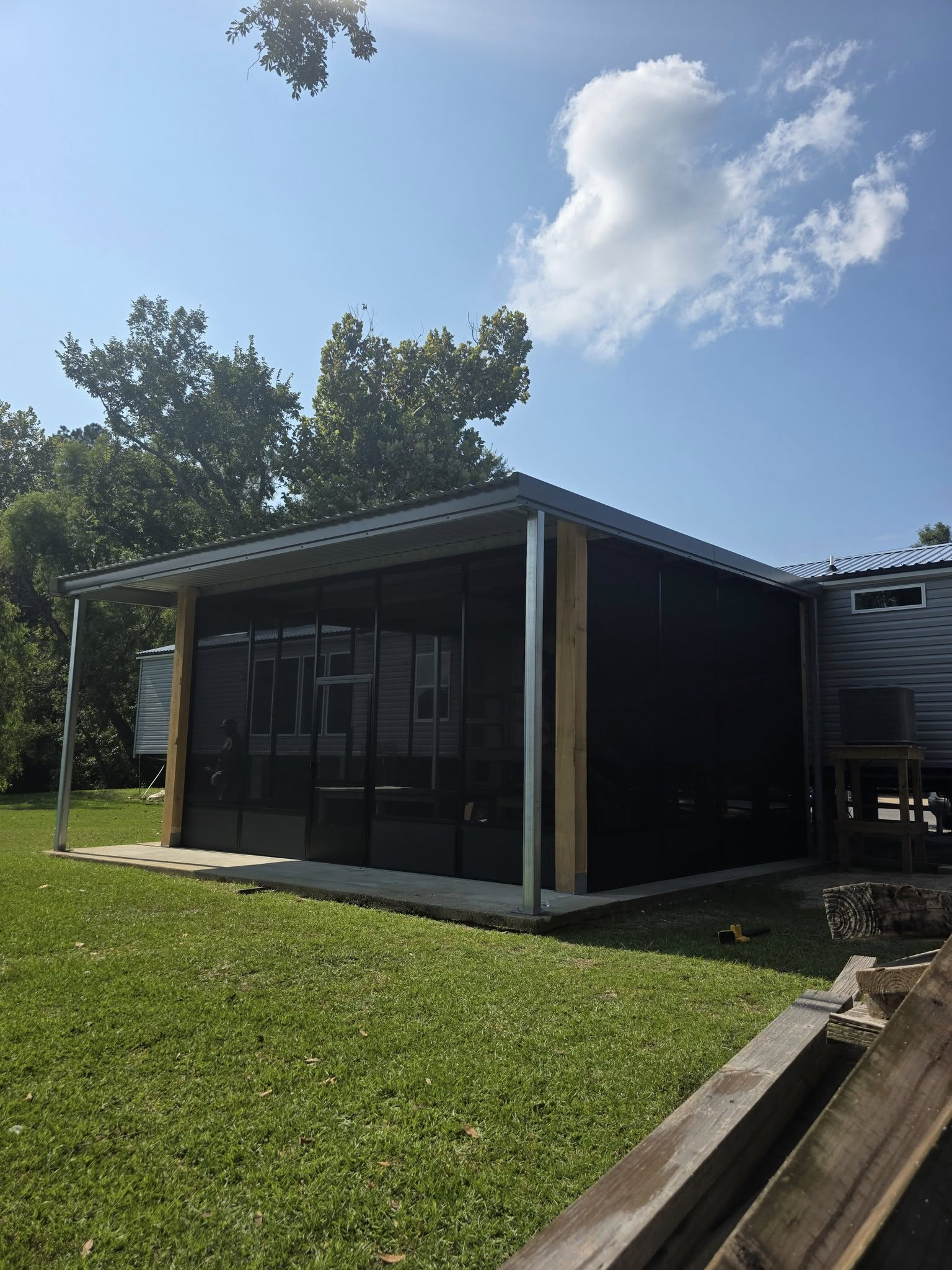 A screened-in porch with a metal roof and wooden beams, sitting on green grass under a blue sky.