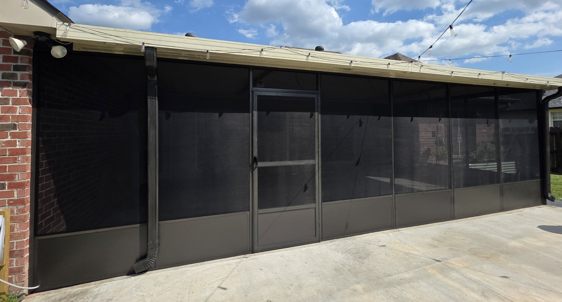 Screened-in patio with dark screens and a door. Brown metal frames on a concrete patio, with brick wall and cloudy sky.