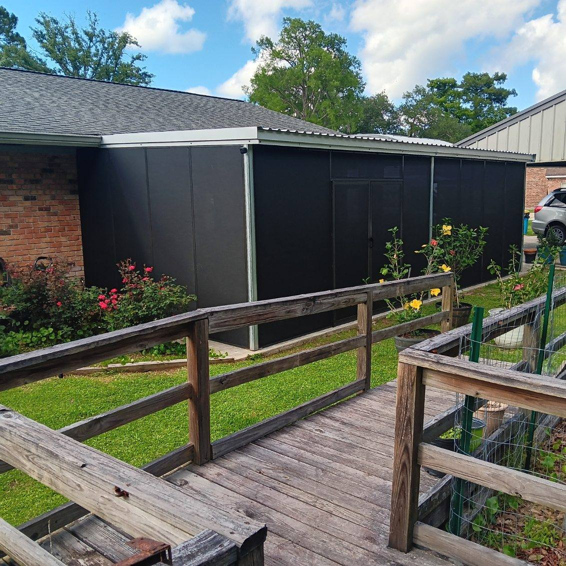 Black shed with ramp and wooden railing; plants in front, brick house to the left.