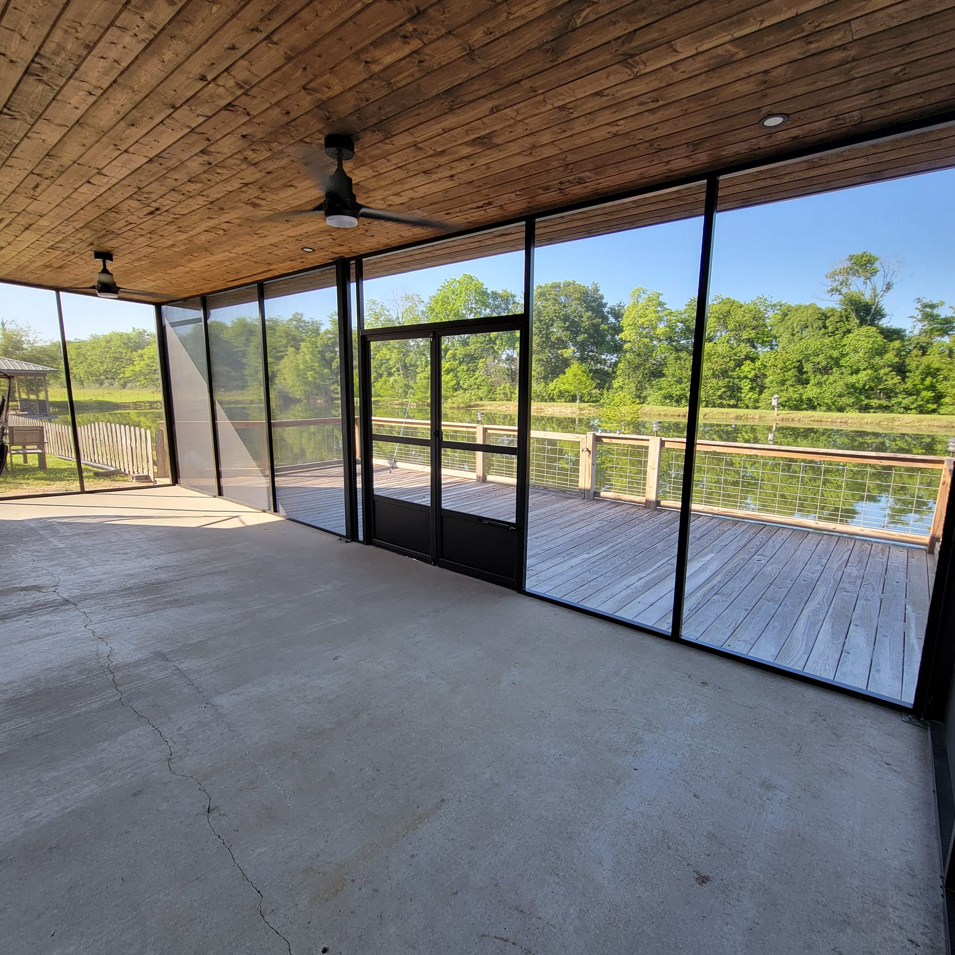 Screened porch overlooking a lake, with a wooden ceiling, concrete floor, and black-framed screen and door.
