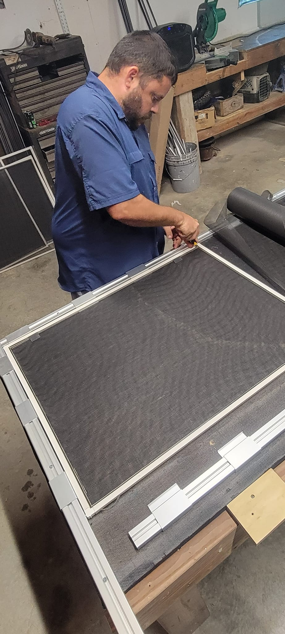 A person working on a screen, indoors. They are using a tool on the frame of a screen, which is lying on a workbench.
