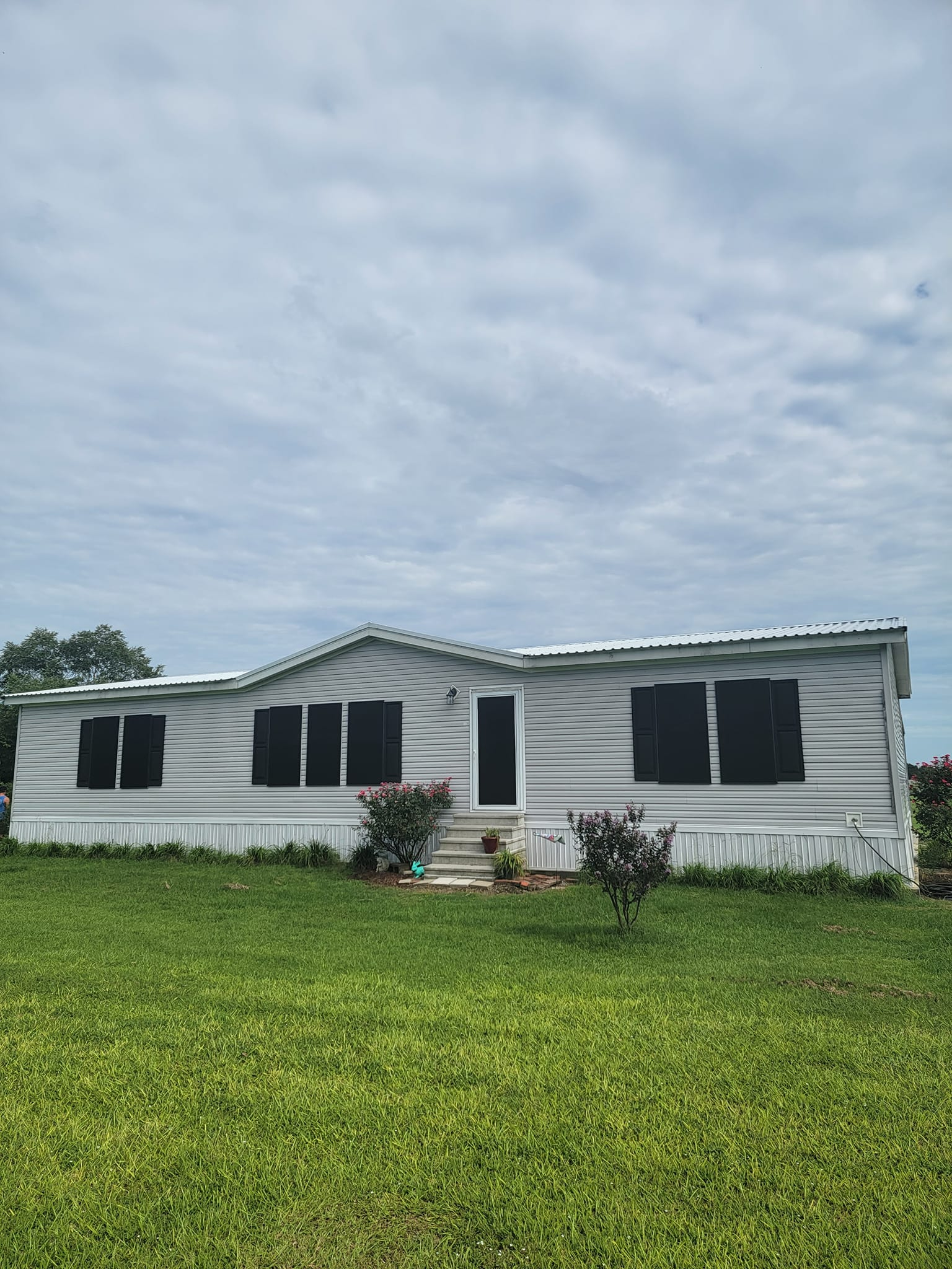 Single-story gray mobile home with black window coverings and a small front yard on a cloudy day.