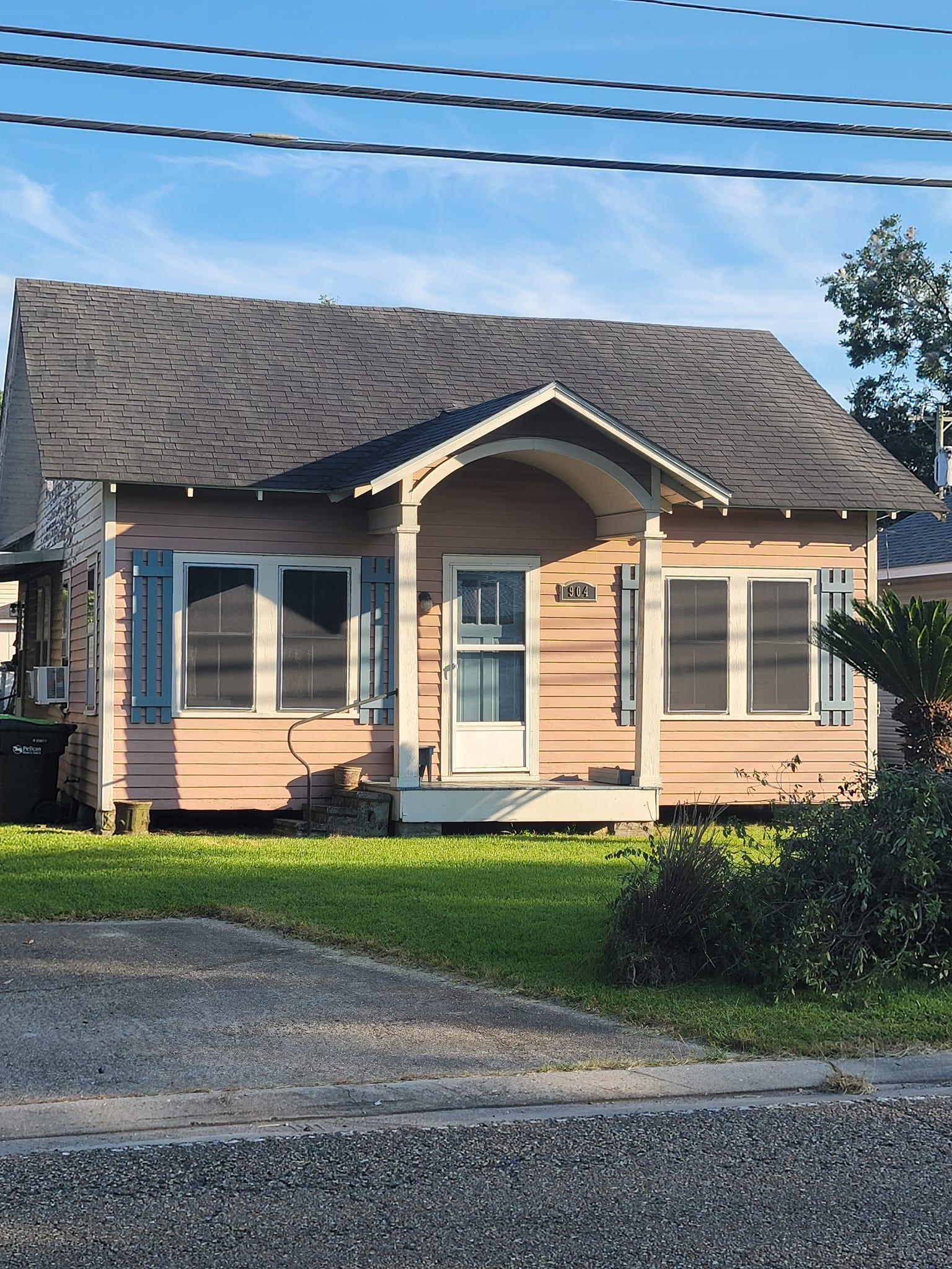 A small, pink house with blue shutters, a porch, and a dark roof, under power lines.