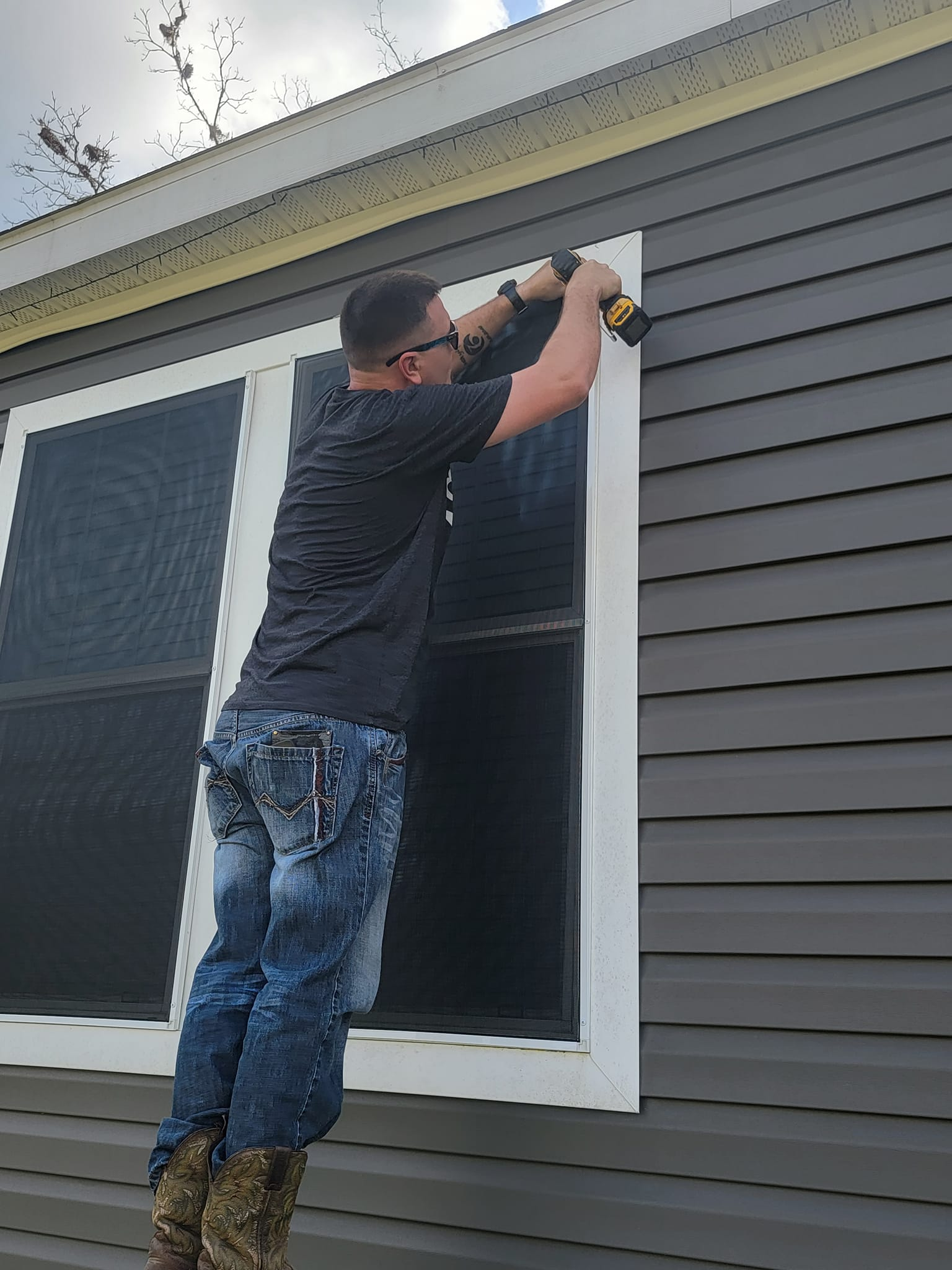 Man on ladder using a drill to work on window trim of a gray house with white trim.