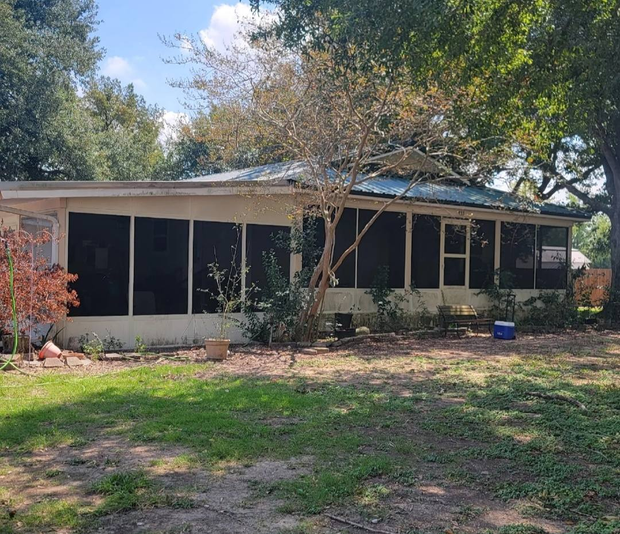 Single-story house with screened porch, surrounded by trees and overgrown lawn, under a partly cloudy sky.