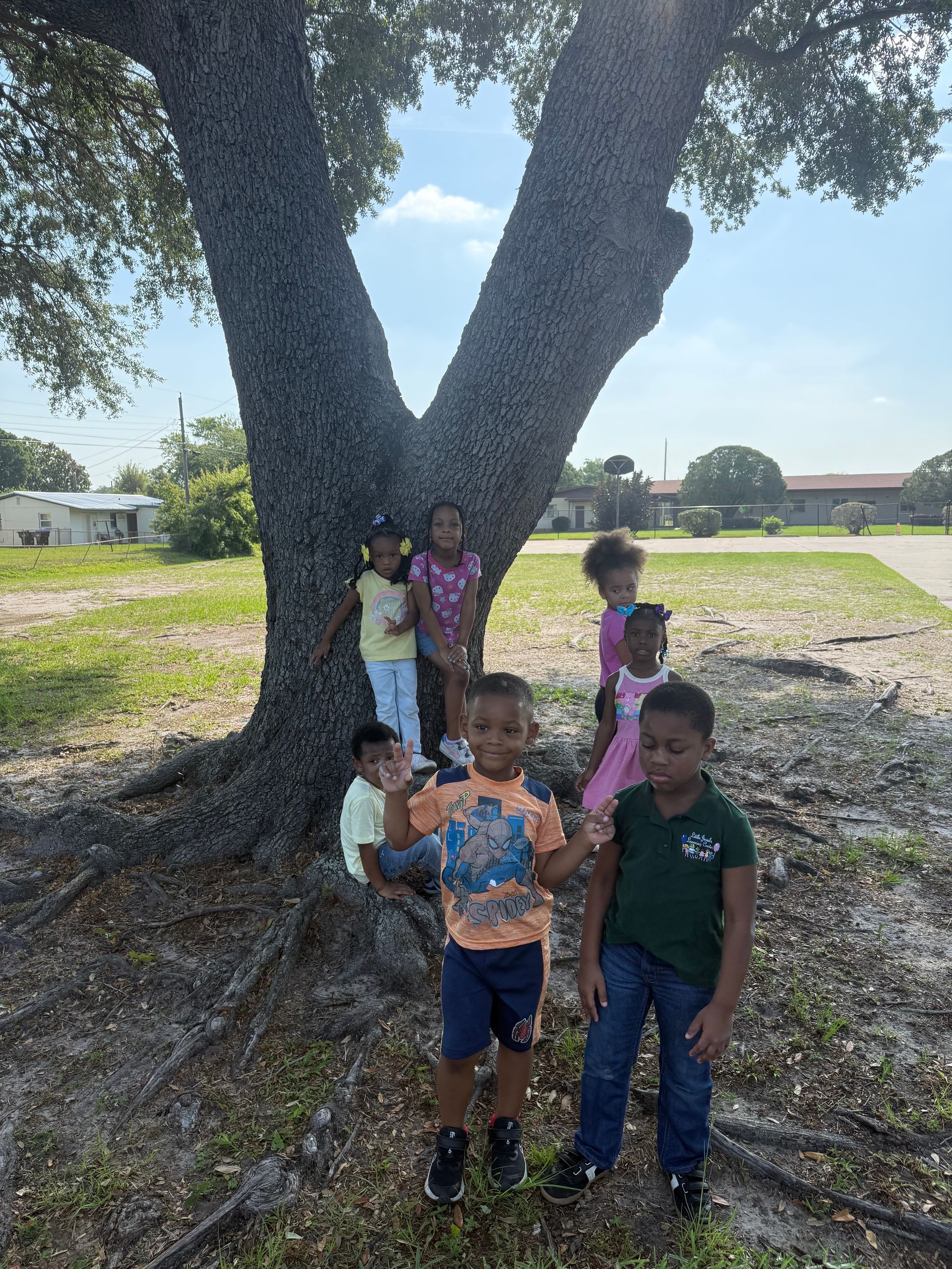Children standing around a large tree outdoors. Some wave, smile. Sunlight, green grass, and houses in the background.